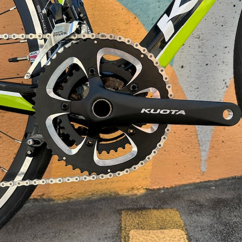 Close-up of a black Kuota bicycle crankset with a chainring and chain, attached to a green and black road bike frame, photographed outdoors on pavement with painted orange and teal markings.